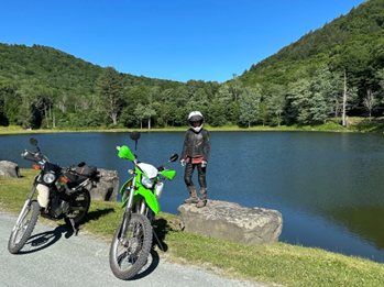 A person standing on a rock next to a lake with two motorcycles in front